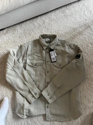 Beige overshirt från C.P. Company - Stilren beige overshirt från C.P. Company i bomull och linne. Klassisk skjortkrage, två bröstfickor med lock och knapp samt knappstängning framtill. Ikonisk linsdetalj på ärmen och raka ärmslut med knappar. Perfekt lager på lager-plagg för en avslappnad look.