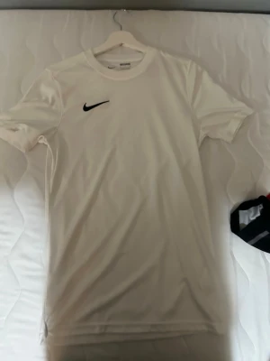 Vit Nike t-shirt i Dri-FIT material - Snygg vit t-shirt från Nike med klassisk svart swoosh-logga på bröstet. Tillverkad i Dri-FIT material som andas och passar perfekt till sport eller vardagsstil. T-shirten har rund halsringning och korta ärmar för en clean och sportig look.