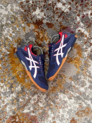 Blå Asics sneakers med vita detaljer - Snygga sneakers från Asics i marinblått med vita och röda detaljer. Skorna har en klassisk låg siluett, meshpaneler för bra ventilation och slitstark gummisula i brunt. Perfekta för dig som gillar sportig stil och vill ha bekväma skor till vardagen.