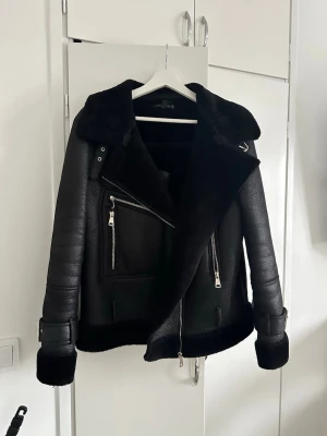 Svart skinnjacka med fuskpäls - Snygg svart skinnjacka med bred krage och fuskpäls på insidan. Jackan har silvriga dragkedjor, två framfickor och spännen vid ärmsluten. Perfekt för dig som vill ha en trendig och skön jacka perfekt till mycket