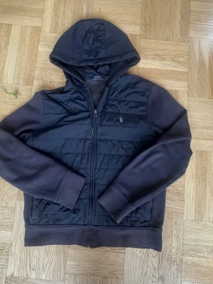  Polo Ralph Lauren cardigan - Snygg mörkblå hoodiejacka från Polo Ralph Lauren med quiltad front, huva och dragkedja. Jackan har en broderad logga på bröstet och ribbade muddar. Perfekt för lager på lager och chill vardagsstil. Storlek M-175.