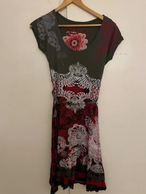 Mönstrad klänning från Desigual - Unik klänning från Desigual med kort ärm, rund halsringning och bälte i midjan. Klänningen har ett färgstarkt mönster i rött, svart, grått och vitt med blommiga detaljer och spetskant nertill. Perfekt för dig som gillar statement-plagg och kreativ design.