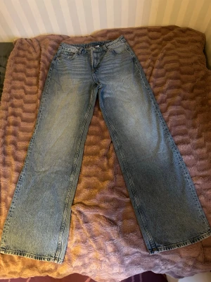 Blåa wide leg jeans från Divided - Säljer ett par ljusblå wide leg jeans från Divided by H&M. Jeansen har klassisk femficksmodell, hög midja och raka, vida ben. Tillverkade i bomull med snygg tvättad look och slitningar för en avslappnad stil.