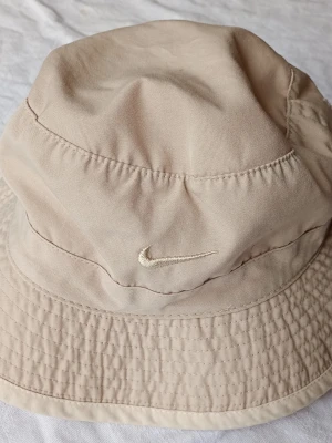 Beige bucket hat från Nike - Säljer en beige bucket hat från Nike med klassisk broderad Swoosh-logga framtill. Hatten är tillverkad i ett mjukt och tåligt bomullstyg och har en avslappnad passform med bred skärm. Perfekt för dig som vill ha en stilren och enkel accessoar.