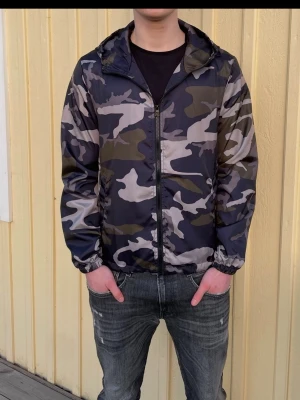 Camo vindjacka med huva - Snygg vindjacka med camouflagemönster i svart, grått och grönt. Jackan har dragkedja framtill, två sidofickor och en praktisk huva. Tillverkad i lätt och smidigt material som passar perfekt för blåsiga dagar.
