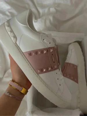 Valentino Garavani sneakers med nitar - Säljer ett par vita Valentino Garavani sneakers i läder med bred rosa rem över foten dekorerad med matchande pyramidformade nitar. Klassisk låg modell med vit sula och subtila detaljer på hälen. Perfekta för dig som vill ha en lyxig och edgy look.