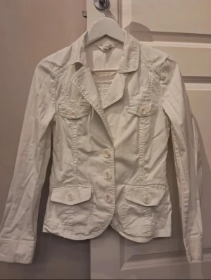 Vit kavaj med fickor och knappar - Stilren vit kavaj med fyra fickor och dekorativa knappar framtill. Klassisk krage och figurnära passform som ger en snygg siluett. Perfekt att matcha till jeans eller kjol för en trendig look.