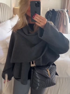 Mörkgrå omlottkappa  - Stilren mörkgrå omlottkappa och stora slag. Kappan har en lös, oversized passform och vida ärmar. Perfekt för lager-på-lager och ger en trendig siluett. Materialet ser ut att vara mjukt och ull-liknande.