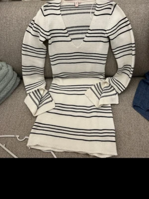 Vit och svart randig stickad klänning - Snygg stickad klänning i vitt med svarta horisontella ränder. Klänningen har v-ringning, lång ärm och lätt utsvängda ärmslut. Passar perfekt för en trendig och avslappnad look.