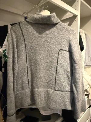 Grå stickad tröja från Zara - Snygg grå stickad tröja från Zara med hög krage och dekorativa svarta sömmar. Tröjan har en avslappnad passform, långa ärmar och ribbstickade muddar. Perfekt för lager på lager och enkel att matcha med olika stilar.