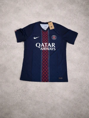 PSG matchtröja Nike mörkblå - Snygg mörkblå PSG matchtröja från Nike med röd och vit detalj i mitten, klubbmärke på bröstet och Qatar Airways-tryck. Tillverkad i lätt och ventilerande material, perfekt för fotboll eller träning. Kortärmad och sportig design.