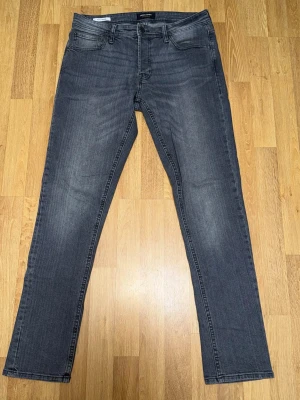 Grå slim fit jeans från Jack & Jones - Snygga grå slim fit jeans från Jack & Jones med klassisk femficksdesign och diskreta slitningar. Jeansen har normal midja, dragkedjegylf och raka ben. Perfekta för en avslappnad och trendig look.