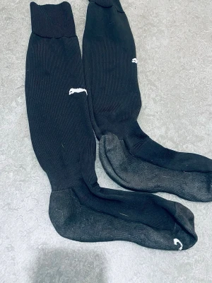 Svarta knästrumpor från Nike - Säljer ett par svarta knästrumpor från Nike med den klassiska vita swoosh-loggan på sidan. Strumporna är ribbade och har märkning för höger och vänster fot. Perfekta till sport eller vardagsoutfits.