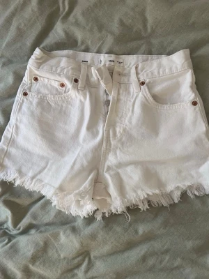 Vita fransiga jeansshorts från Mango - Säljer ett par vita jeansshorts från Mango med fransig kant nertill. Shortsen har fem fickor, bälteshällor och knappgylf. Perfekta för varma dagar och har en klassisk, avslappnad look.