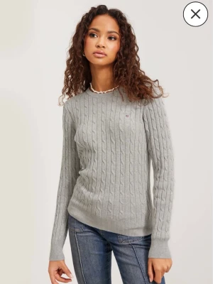 Ljusgrå kabelstickad tröja Gant - Snygg ljusgrå kabelstickad tröja från Gant med rund halsringning och diskret logga på bröstet. Tröjan har långa ärmar och ribbade muddar vid ärmslut och nederkant. Perfekt till jeans för en clean look.