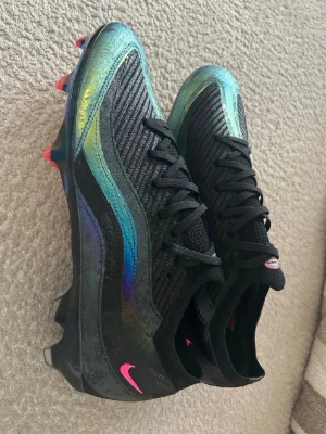 Nike Air max 95 Mercurial Vapor fotbollskor  - Säljer ett par Nike Mercurial Vapor fotbollsskor i svart med skimrande blå och gröna detaljer. Skorna har rosa Nike-logga och en färgglad yttersula i orange, gult och blått. Storlek 44 men passar 45 även. 10/10 skick! Priset kan diskuteras!