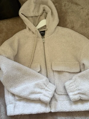 Beige teddyjacka med huva från ONLY - Mysig beige teddyjacka från ONLY med dragkedja framtill, två stora fickor med lock och huva med dragsko. Har aldrig använt 