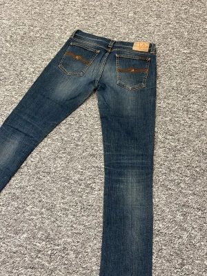 Blå jeans från Nudie Jeans - Snygga blå jeans från Nudie Jeans med klassisk femficksdesign och orange broderi på bakfickorna. Jeansen har en smal passform och är tillverkade i denim med lätt slitning för en cool look.
