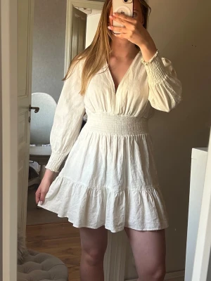 Vit kort klänning med v-ringning - Superfin vit kort klänning med v-ringning och långa ärmar. Klänningen har smock i midjan och vid ärmsluten, samt volang nertill för en extra söt look. Tillverkad i ett lätt och luftigt material, perfekt för varmare dagar. Säljer pga att den är förliten. Perfekt till skolavslutning mm.