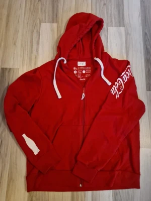 Limiterad Röd Coca-Cola hoodie med dragkedja - Limited Edition Tröja endast 1000 tröjor gjordes! Och den här kan bli din ! Cool röd hoodie från Coca-Cola med vit logga på ärmen och vit flask-symbol på andra ärmen. Hoodien har huva med vita snören och dragkedja framtill. Perfekt för dig som gillar retrostil och ikoniska märken. Materialet är mjukt och bekvämt, troligen bomullsblandning.