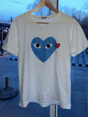 Vit Comme des Garçons Play t-shirt - Vit t-shirt från Comme des Garçons Play med ett stort blått hjärta och ögon på bröstet samt ett litet rött hjärta bredvid. Klassisk rund hals och korta ärmar. Perfekt för dig som gillar streetwear och vill sticka ut lite extra.