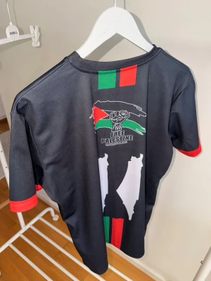 Svart Free Palestine t-shirt  - Svart t-shirt med tryck i rött, grönt och vitt samt texten 'Free Palestine' på ryggen. T-shirten har korta ärmar med röda detaljer och är tillverkad i ett lätt och syntetiskt material. Perfekt för dig som vill visa ditt stöd och ha en unik stil storlek XL menar passar mer som L 