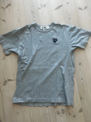 Grå t-shirt från Comme des Garçons Play - Grå t-shirt från Comme des Garçons Play med det ikoniska svarta hjärta-loggan på bröstet. Klassisk passform med rund hals och korta ärmar.  T-shirten är i mycket gott skick utan hål och fläckar. Storleken är L men passar M då det är japanska mått