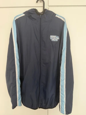 Blå vindjacka Norra Läktaren Malmö - Snygg marinblå vindjacka med huva och dragkedja framtill. Jackan har ljusblå och vita ränder längs ärmarna samt ett stort Norra Läktaren Malmö-tryck på bröstet. Perfekt för dig som vill visa ditt stöd på läktaren. Lätt och smidig att bära.