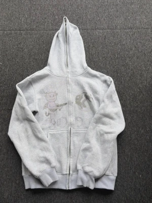 Aim for the moon - Säljer en ljusgrå hoodie från aim for the moon med dragkedja och huva. Framsidan har ett coolt tryck med en tecknad figur och detaljer i svart och grått. Hoodien har två fickor fram och ribbade muddar. Perfekt för en avslappnad streetstyle-look.