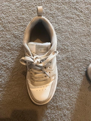 Vita Champion sneakers i läder - Snygga vita sneakers från Champion med klassisk låg profil. Skorna har ovandel i läder och textil, vita snören och diskret logga på plösen. Yttersulan är i gummi med Champion-mönster undertill. Perfekta för en clean och sportig look.