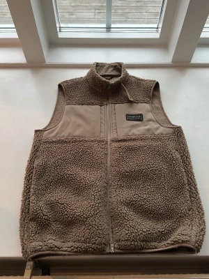 Beige teddyväst från Barbour - Säljer en beige teddyväst från Barbour International med hög krage och dragkedja framtill. Västen har en mjuk, fluffig yta i teddy och släta partier upptill. Perfekt för lager på lager och riktigt snygg till höstlooken.