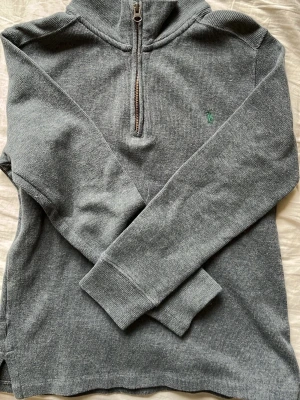 Grå halvzip från Polo Ralph Lauren (M för barn) - Snygg grå tröja från Polo Ralph Lauren med halvzip och hög krage. Tröjan har ett diskret grönt logobroderi på bröstet och är tillverkad i mjukt bomullsmaterial. Perfekt för lager på lager och enkel att matcha med olika outfits.