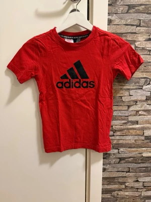 Röd Adidas t-shirt med logga - Röd t-shirt från Adidas med klassisk svart logga framtill. T-shirten har rund halsringning och korta ärmar. Tillverkad i mjuk bomull som är skön att bära. Perfekt för en sportig och avslappnad stil.