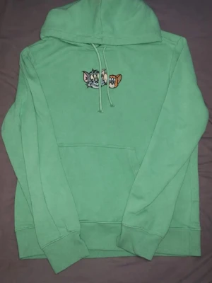 Grön Tom & Jerry hoodie - Snygg grön hoodie med broderat Tom & Jerry-motiv på bröstet. Klassisk modell med huva och dragsnören samt stor magficka. Perfekt för dig som gillar tecknade serier och vill ha en chill och lekfull stil.