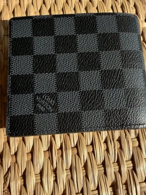 Plånbok i rutat mönster - Snygg plånbok i klassiskt rutat mönster. Plånboken är svart och grå och är tillverkad i tåligt läder. Perfekt för dig som gillar stilrena accessoarer med lyxig känsla.