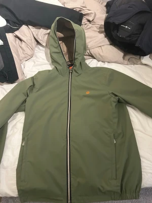 Vindjacka - Sail Racing - Snygg olivgrön vindjacka från Sail Racing med huva och hel dragkedja framtill. Jackan har två sidofickor, elastiska muddar och diskret logga på bröstet. Materialet är lätt och vattenavvisande, perfekt för blåsiga och kalla förhållande