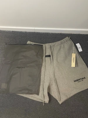 Grå shorts från Essentials - Snygga grå shorts från Essentials med elastisk midja och snörning. Shortsen har en avslappnad passform och är tillverkade i mjuk bomull. Perfekta för sommardagar och chill outfits. Essentials-logga tryckt på ena benet. Levereras med originalpåse.
