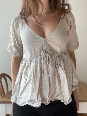 Beige blus med knyt och v-ringning - Supersöt beige blus med v-ringning och knyt i midjan. Puffiga korta ärmar och avslappnad passform ger en romantisk vibe. Tillverkad i ett lätt och luftigt material, perfekt för varmare dagar.