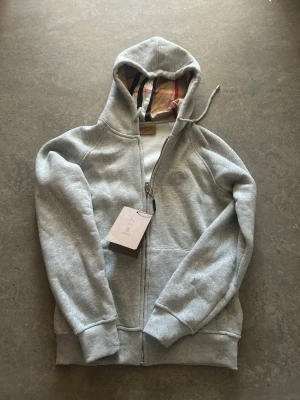 Grå zip hoodie från Burberry - Snygg grå zip hoodie från Burberry med klassiskt rutigt foder i huvan och dragsko. Tillverkad i mjuk bomullsblandning och har ribbade muddar samt fickor framtill. Perfekt för dig som vill ha en stilren och exklusiv hoodie med ikoniska Burberry-detaljer.