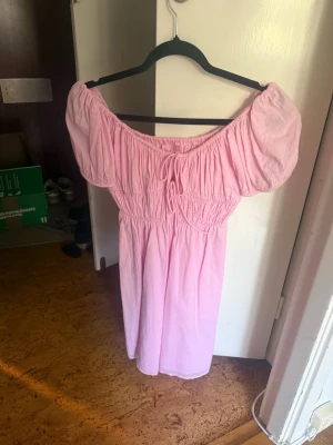 Ljusrosa offshoulder klänning - Söt ljusrosa klänning med offshoulder-modell och puffiga korta ärmar. Klänningen har resår i midjan och en liten knytning framtill. Perfekt för varma dagar och ger en romantisk look.