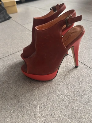 Bruna peep-toe pumps från Scorett - Säljer ett par bruna pumps från Scorett med öppen tå och hög platåklack. Skorna har en snygg slingback-rem med spänne och är tillverkade i läder. Perfekta för dig som vill ha ett statement-plagg till din outfit.
