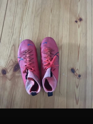 Röda Nike Mercurial fotbollsskor - Säljer ett par röda Nike Mercurial fotbollsskor med högt skaft och svarta detaljer. Skorna har snörning och dobbar för bra grepp på planen. Tillverkade i syntetmaterial som ger en lätt och smidig känsla. Perfekta för dig som vill ha snabbhet och kontroll på fotbollsplanen.
