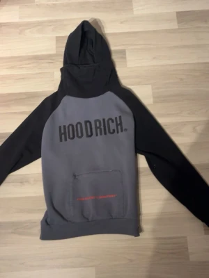  Hoodrich hoodie  - Säljer en grå och svart hoodie från Hoodrich med stor logga på bröstet och ficka framtill. Hoodien har en rymlig huva med ventilationshål och är tillverkad i mjukt bomullsmaterial. Perfekt för dig som gillar streetwear och vill ha en cool look.