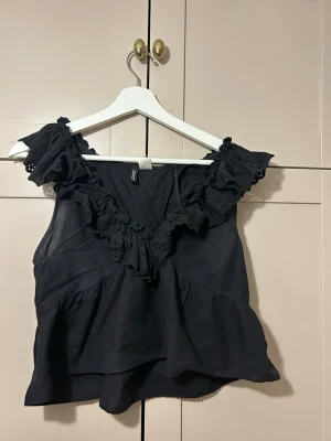 svart volangblus med spetsdetaljer - Säljer en svart blus från H&M med volang och spetsdetaljer runt halsen. Blusen är ärmlös och har en luftig passform som ger en cool och trendig look.