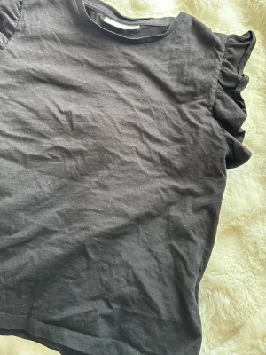 Svart volangtopp från Gina Tricot - Snygg svart topp från Gina Tricot med volangdetaljer på axlarna. Tillverkad i mjuk bomull och har en klassisk rund halsringning. Perfekt för dig som vill ha en enkel men trendig topp till garderoben.