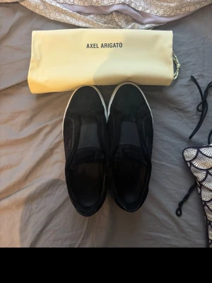 Svarta sneakers från Axel Arigato - Stilrena svarta sneakers från Axel Arigato med vit sula och diskret guldfärgad logga på sidan. Skorna är i mocka och har en minimalistisk design utan snörning, vilket ger en clean look. Perfekta för dig som gillar enkel och modern stil. Har ett hål i undersidan men inget som kommer in i själva skorn!