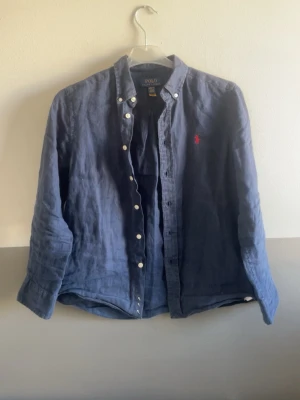 Blå linneskjorta från Polo Ralph Lauren - Snygg blå skjorta i linne från Polo Ralph Lauren med klassisk krage och knappar framtill. Skjortan har lång ärm och den ikoniska röda loggan broderad på bröstet. Perfekt för en avslappnad men stilren look.