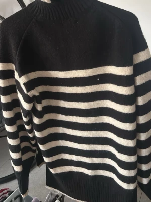 Svartvit randig stickad tröja - Säljer en svart stickad tröja med vita horisontella ränder. Tröjan har hög hals, långa ärmar och ribbstickade muddar. Passar perfekt för dig som gillar klassisk och enkel stil. Materialet ser ut att vara mjukt och bekvämt.