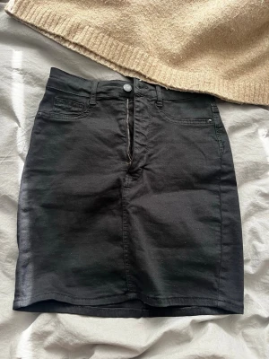 Svart jeanskjol med fickor - Svart jeanskjol med klassisk femficksdesign, dragkedja och knapp framtill. Kjolen är rak i modellen och slutar ovanför knäna. Perfekt att matcha med en stickad tröja eller t-shirt.