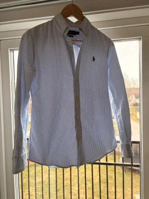 Randig skjorta från Polo Ralph Lauren - Säljer en klassisk långärmad skjorta från Polo Ralph Lauren med smal passform. Skjortan är vit och ljusblå med tunna ränder, har knappar framtill och broderad logga på bröstet. Perfekt för dig som gillar stilrena plagg med ikonisk design.                             Säljer skjortan eftersom den blivit för liten för mig. Grymt skick då den bara är använd fåtal gånger. Skjortan är i storlek S och är slim fit vilket gör den lite tightare än en vanlig S